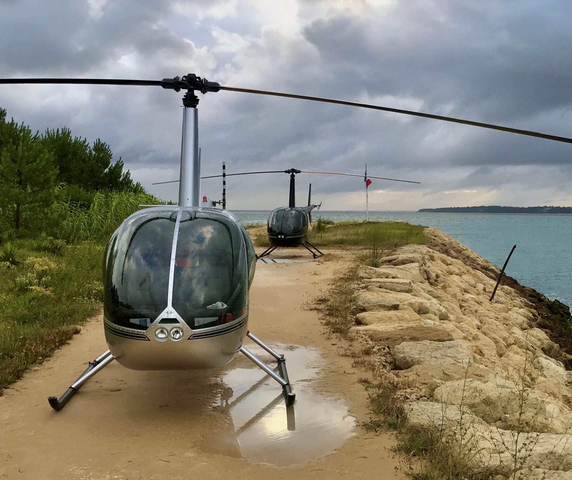 2 x Robinson R44 helicopters landing on a narrow strip of ground next to the sea