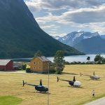 Helipaddy helicopters parked on scenic field with mountain and lake views, near a vibrant yellow house.