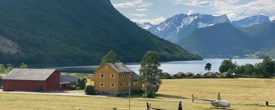 Helipaddy helicopters parked on scenic field with mountain and lake views, near a vibrant yellow house.