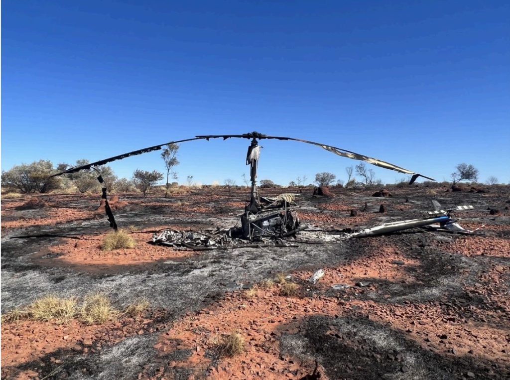 Charred helicopter remains in a desolate landscape; Helipaddy incident site under clear blue sky.