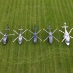 Helipaddy: Eight helicopters aligned on a grassy field, showcasing diverse rotors and designs from an aerial view.