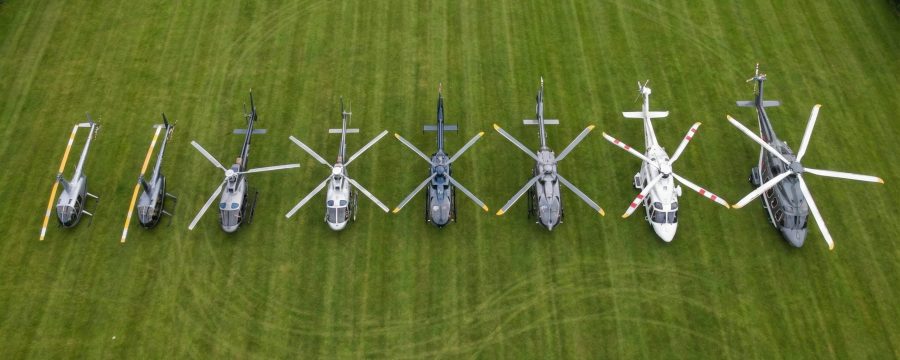 Helipaddy: Eight helicopters aligned on a grassy field, showcasing diverse rotors and designs from an aerial view.