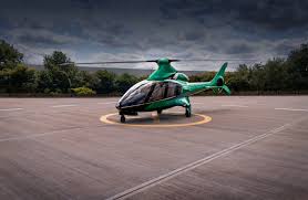 Green Hill helicopter on Helipaddy helipad, surrounded by trees, under a cloudy sky.