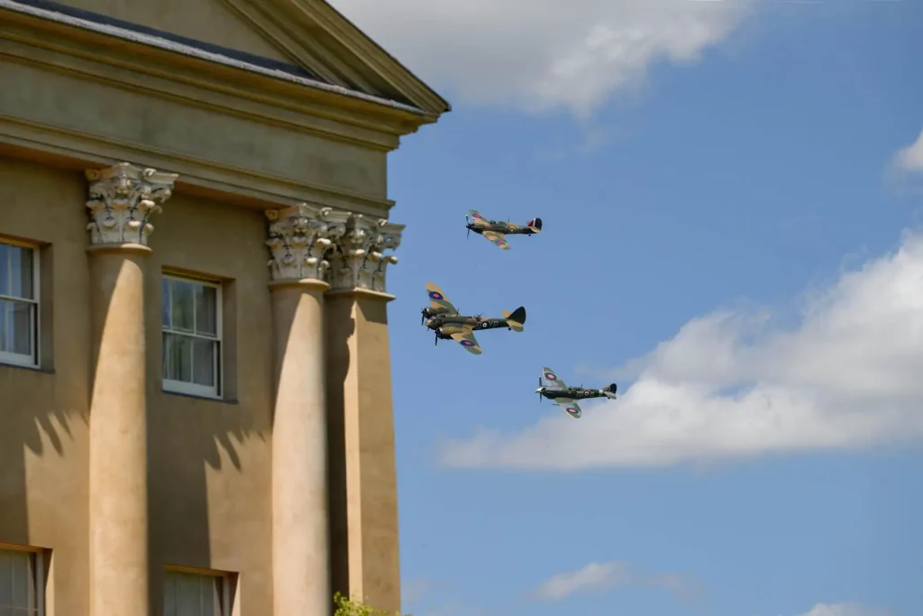 Historic planes soar near a stately building under a blue sky, showcasing Helipaddy at its best.